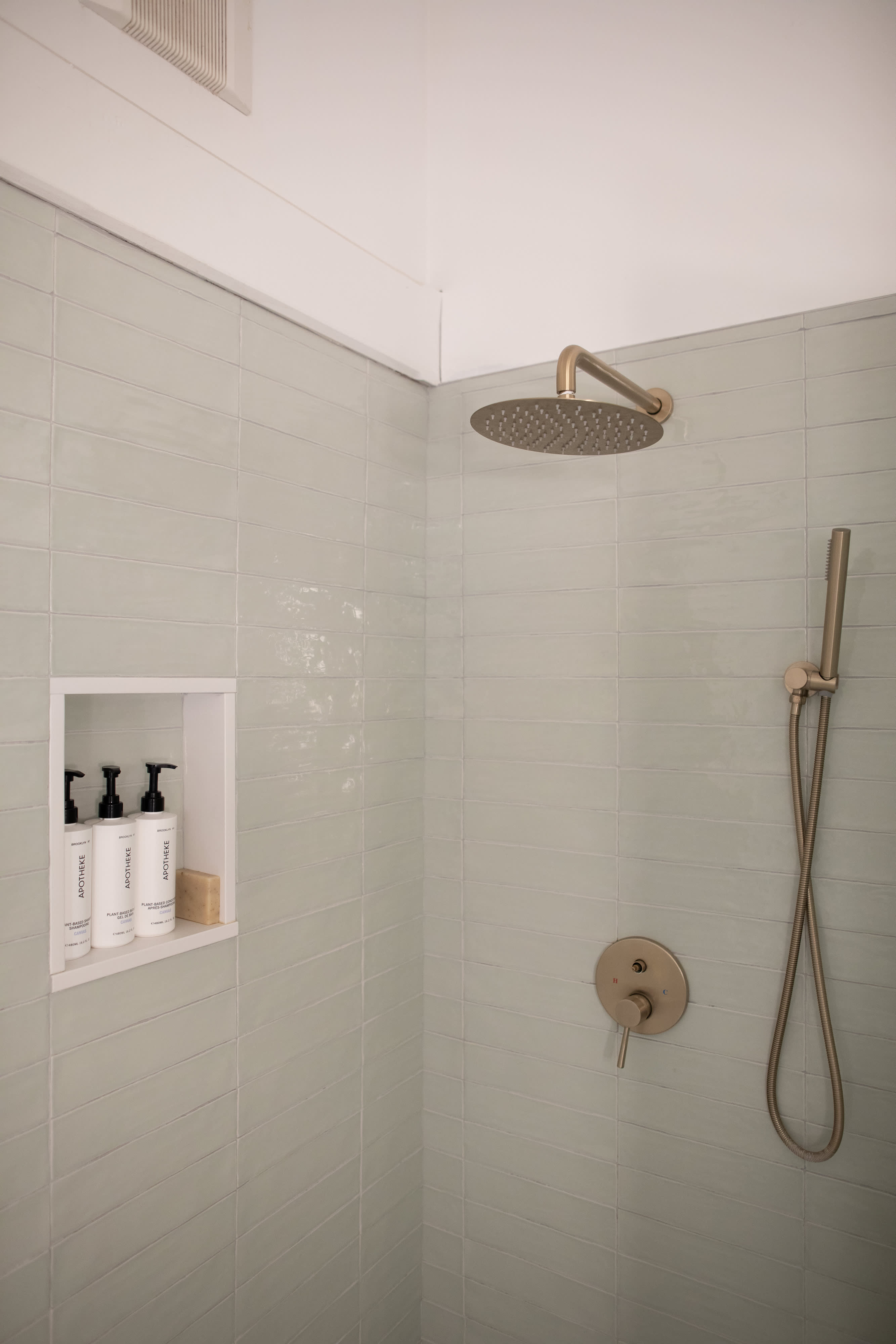 Modern shower with light green subway tiles, a built-in niche holding toiletries, and brushed gold fixtures including a rain shower head and a handheld sprayer.