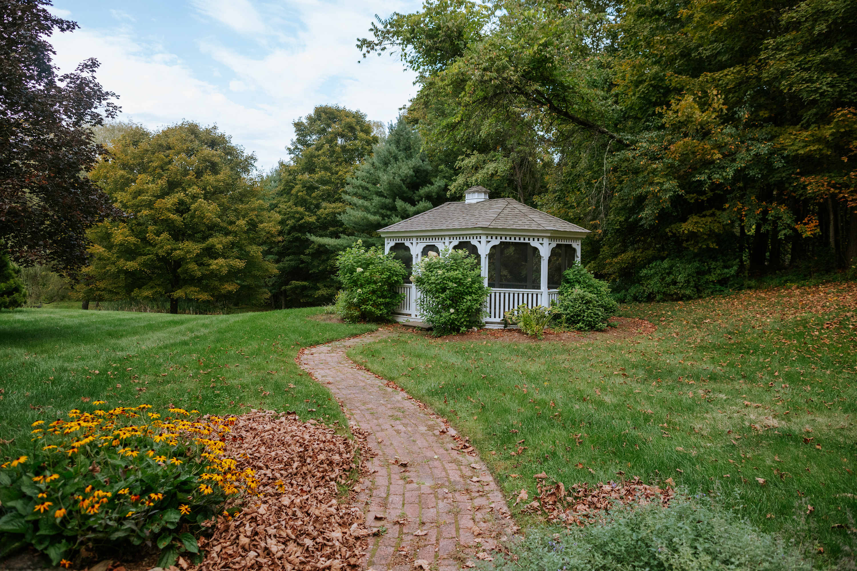 A charming white gazebo with a screened enclosure sits amidst a vibrant green lawn, accessible by a winding brick pathway. The surrounding landscape is dotted with mature trees and shrubs, some showing autumn colors.