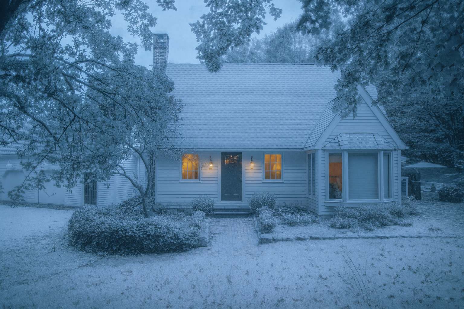 The inviting front entrance of a white house, softly covered in a fresh layer of snow, with warm lights illuminating the windows and front door. Snow-dusted bare tree branches frame the scene, enhancing the winter charm.