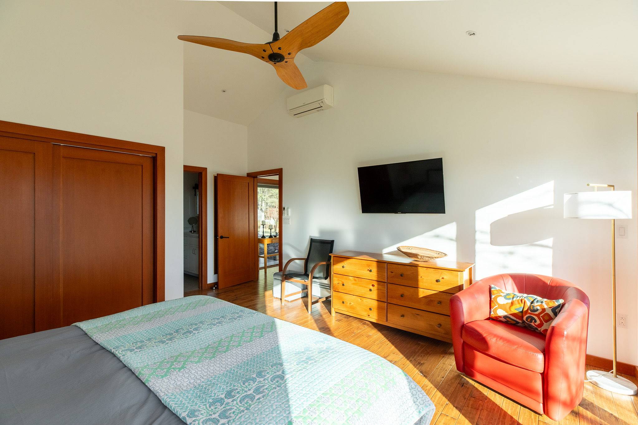 Another perspective of the bright bedroom, showcasing a wooden dresser with a wall-mounted television, a vibrant red armchair, and a glimpse into an adjacent hallway with warm wooden doors.