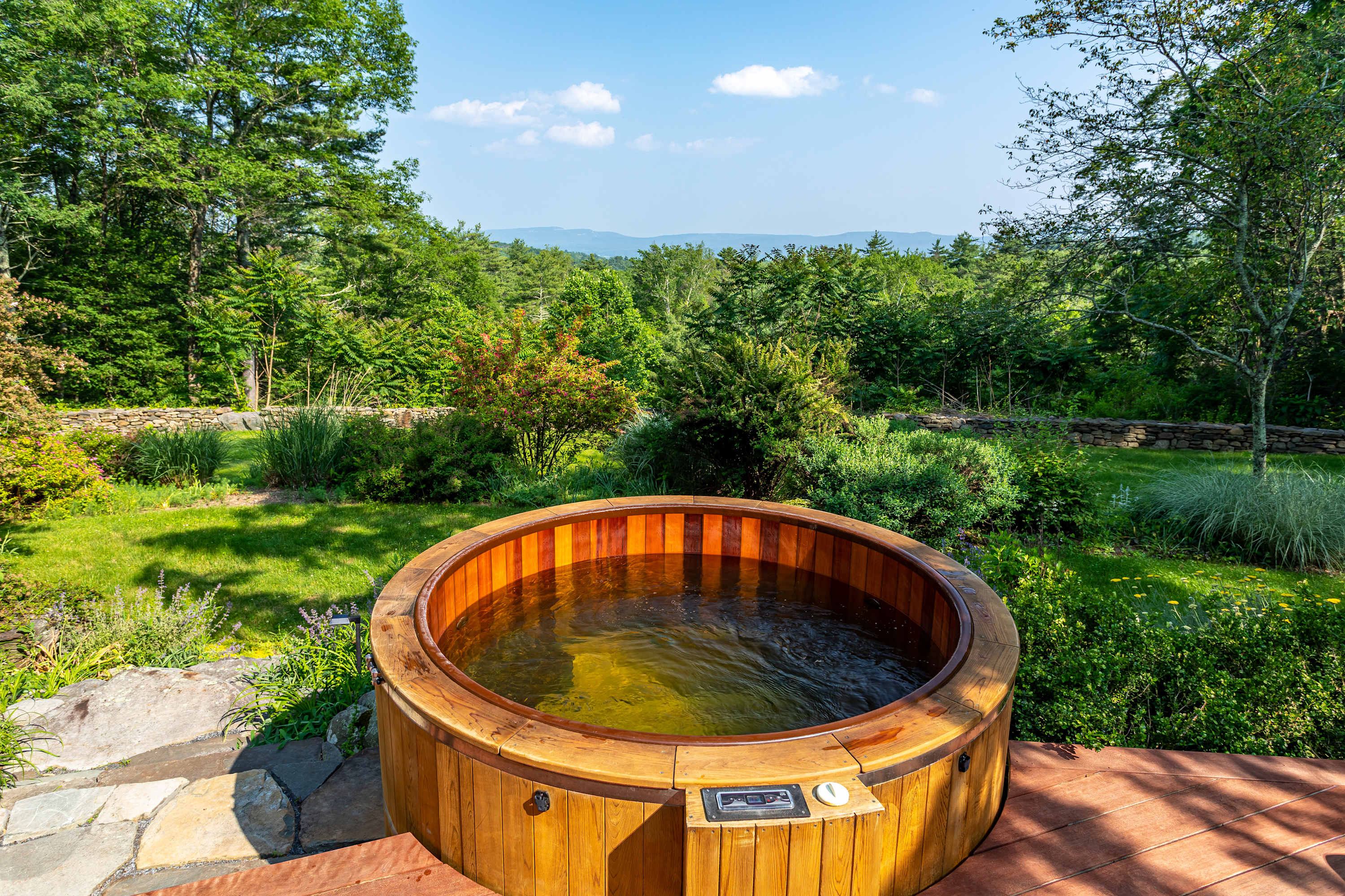 A close-up view of a round wooden hot tub filled with water, set into a wooden deck. The surrounding landscape features a manicured green lawn, lush bushes, and distant trees under a bright sky.