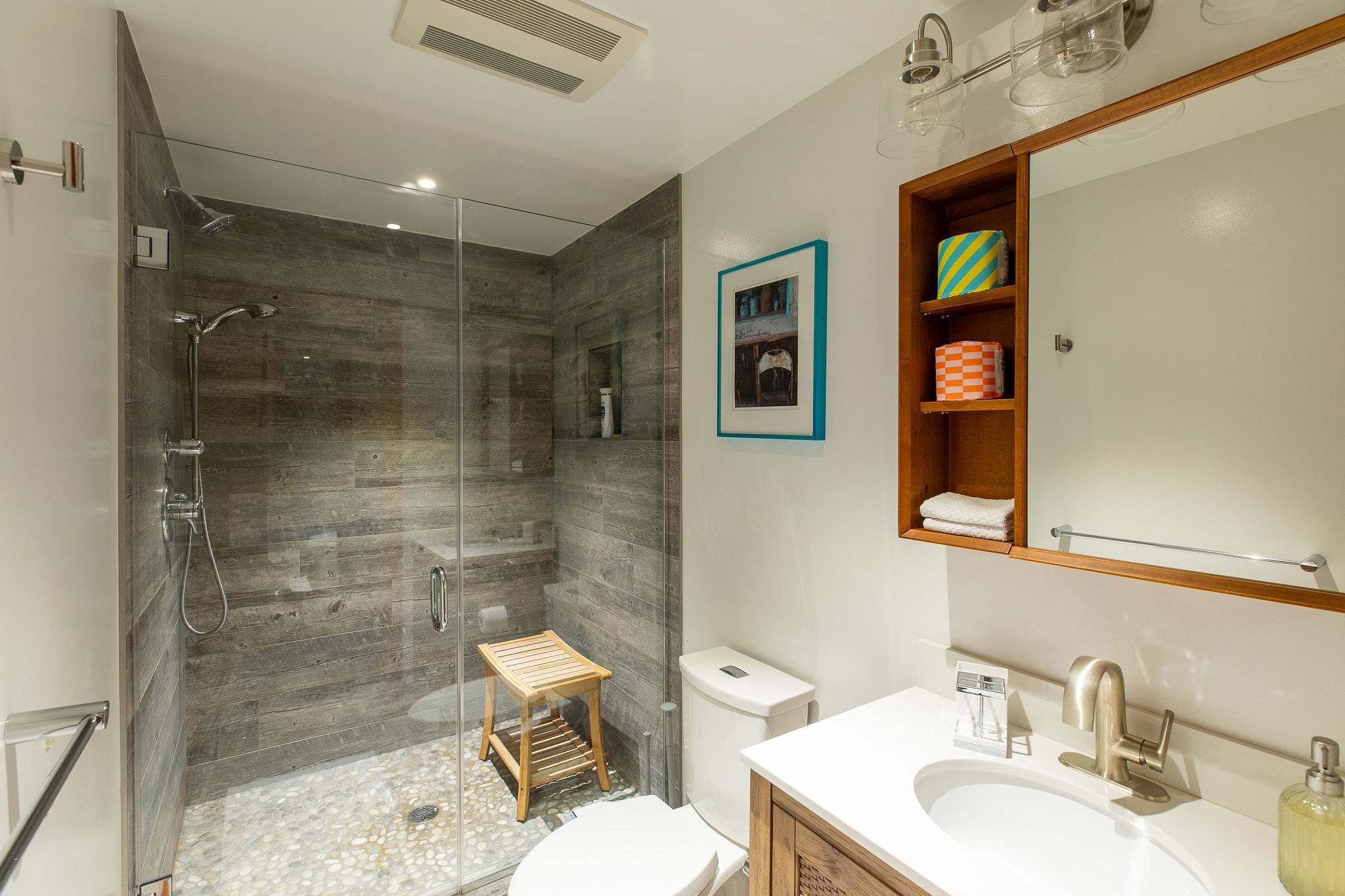 A modern bathroom with a walk-in shower featuring wood-look tile walls and a pebble floor, a white vanity with a sink, a toilet, and a wooden medicine cabinet with a mirror.