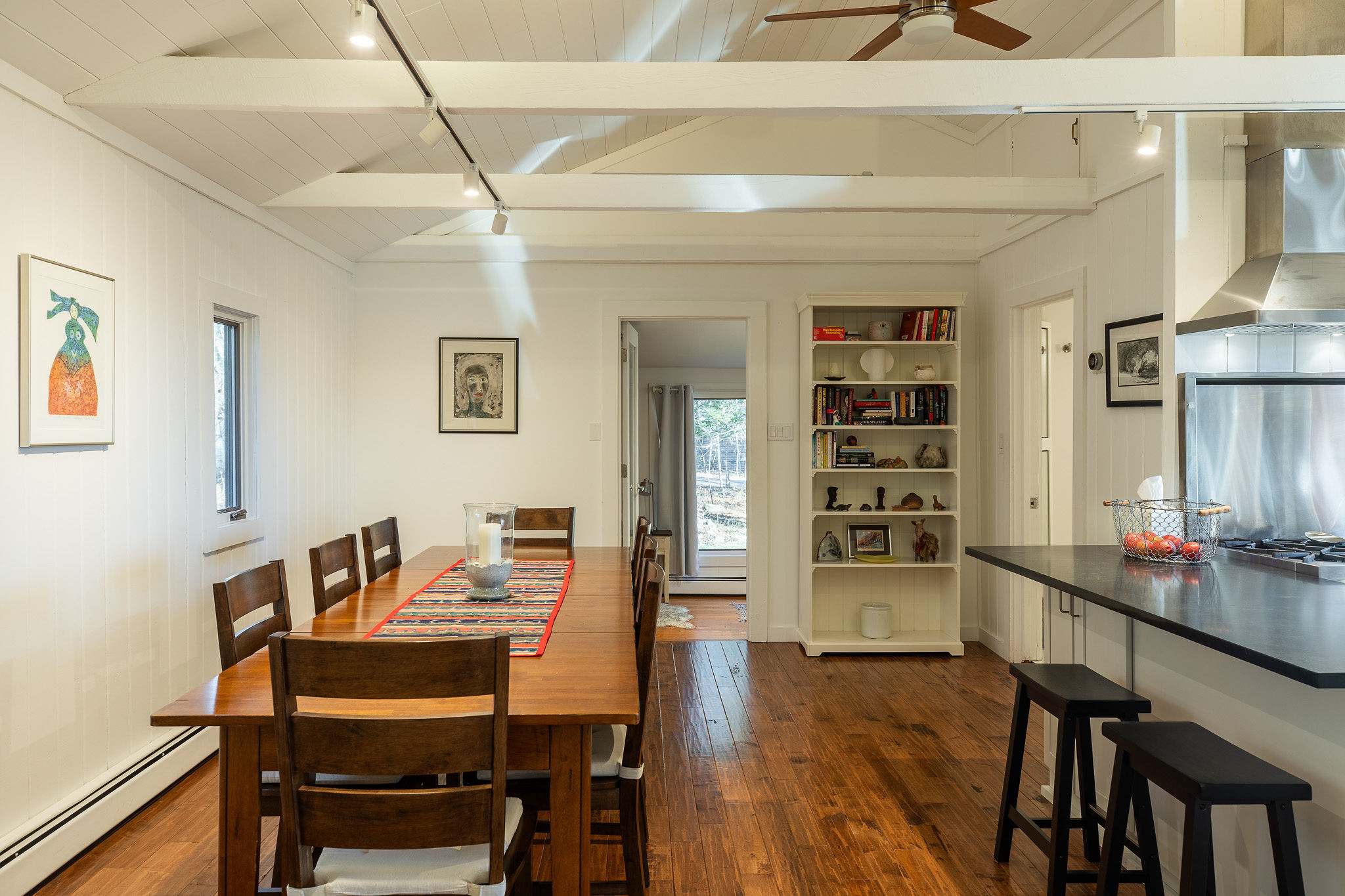 A spacious dining area with a long wooden table set for eight, complemented by matching chairs, rich hardwood floors, and a white paneled vaulted ceiling with exposed beams and track lighting.