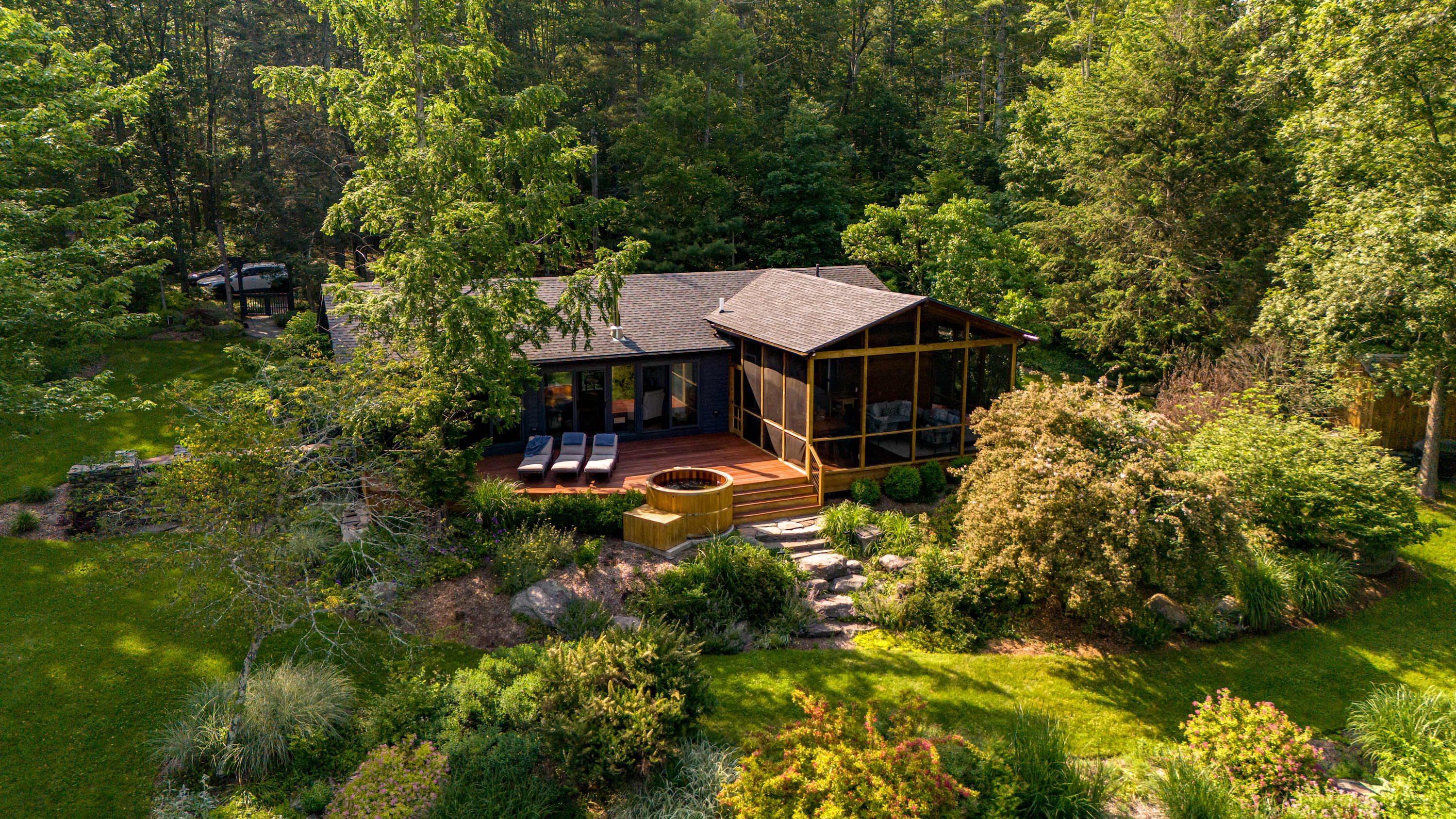 Aerial view of a dark-sided house nestled among lush green trees, featuring a large wooden deck with a hot tub, a screened porch, and well-maintained landscaping.