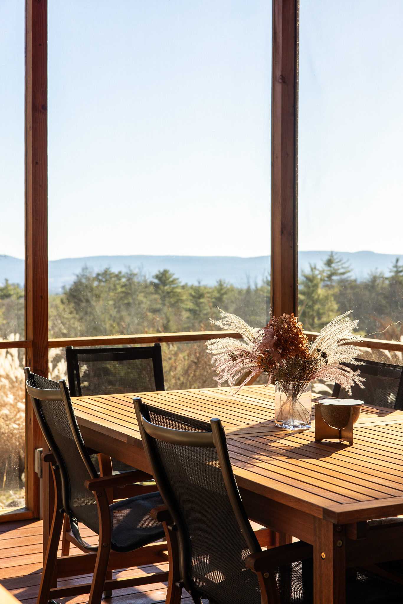 A bright screened-in porch features a slatted wooden dining table with four black mesh chairs, set against a backdrop of distant mountains and a sunny, wooded landscape. A vase with dried floral arrangements sits on the table.