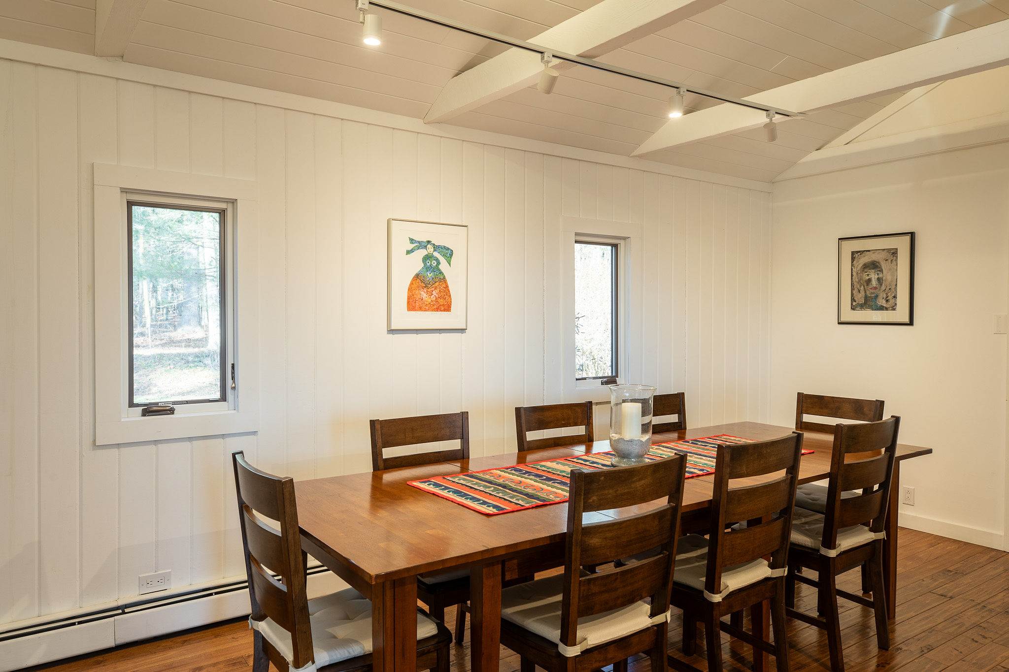 A well-lit dining area featuring a long wooden table with matching chairs, warm hardwood floors, and white paneled walls adorned with two windows that provide views of the surrounding trees.