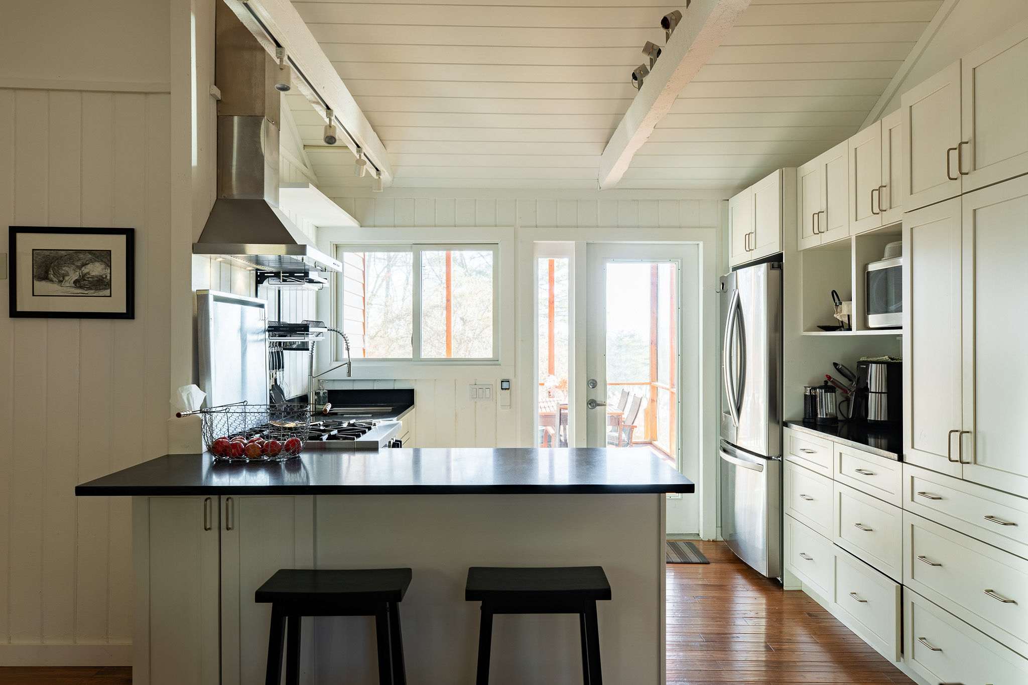 A bright kitchen with white cabinetry, dark countertops, a gas range with a prominent stainless steel hood, and a large window above the sink, providing natural light and a view, with access to an outdoor deck.