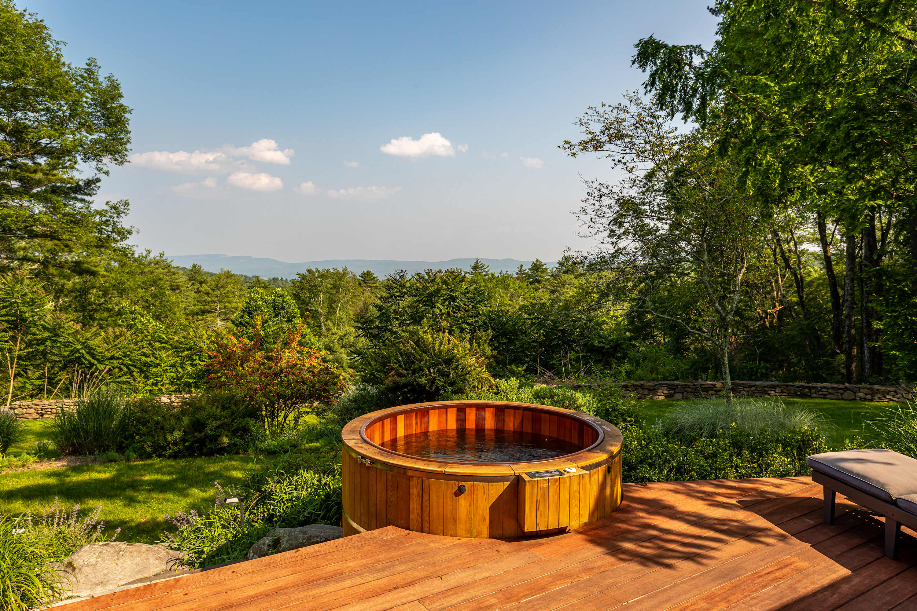 A round wooden hot tub is set into a spacious wooden deck, surrounded by lush green trees and a distant mountain range under a clear blue sky. The hot tub water is visible and appears to be bubbling.