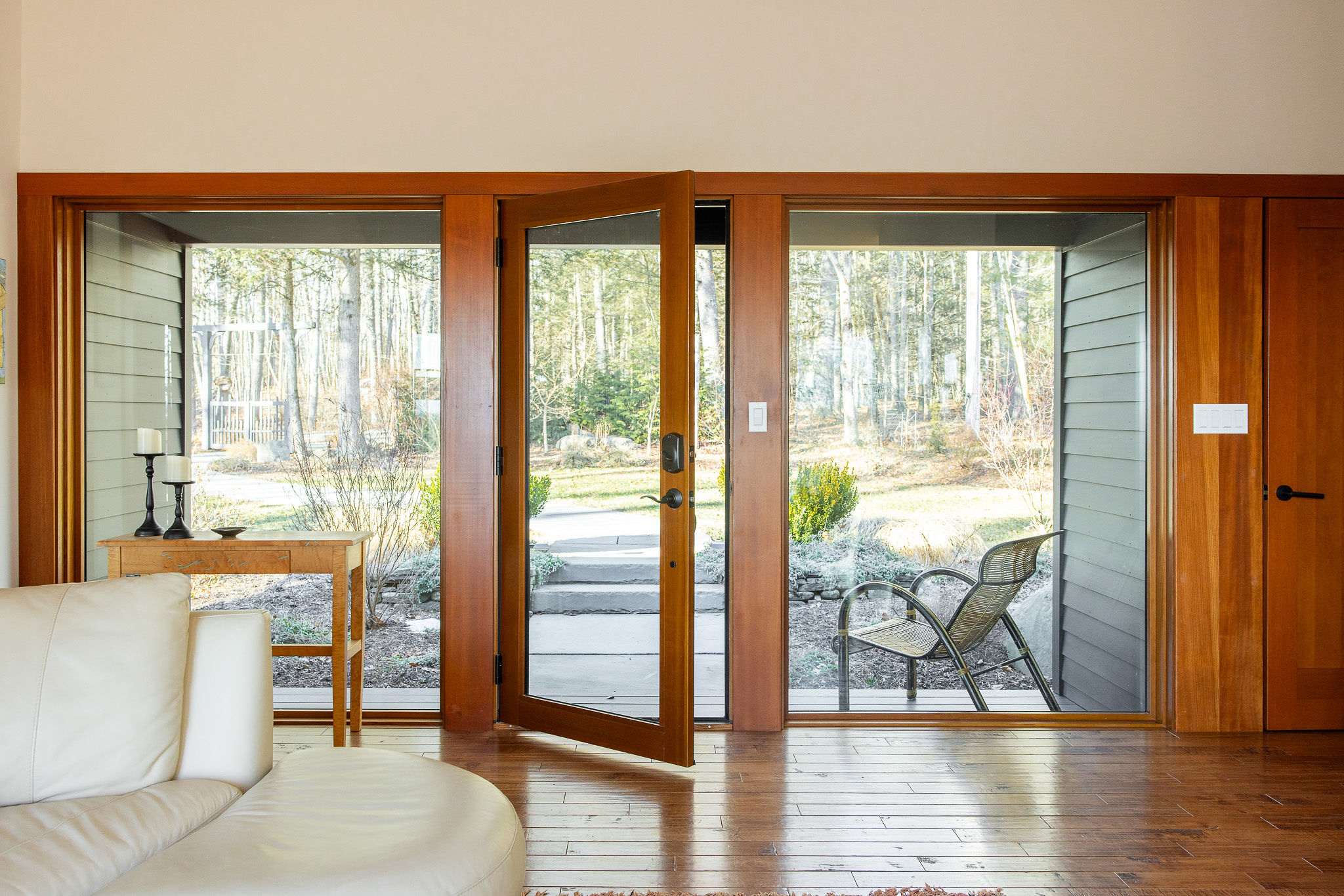 A bright living space with warm hardwood floors, a plush white sofa, and large wooden-framed glass doors that open to an outdoor path and a serene wooded landscape.