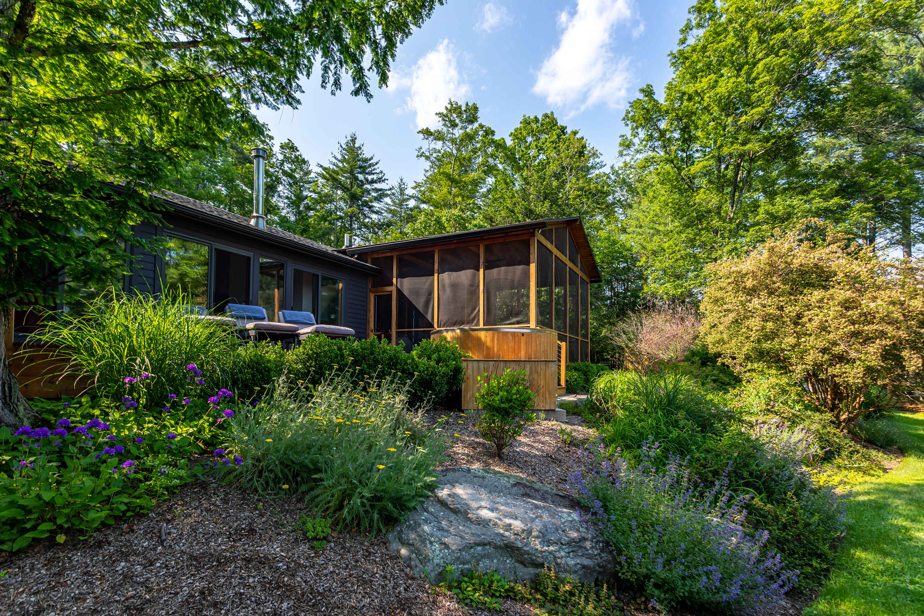 Exterior view of the house from a landscaped garden, showcasing a wooden deck with lounge chairs and a screened porch, surrounded by vibrant greenery and large trees.
