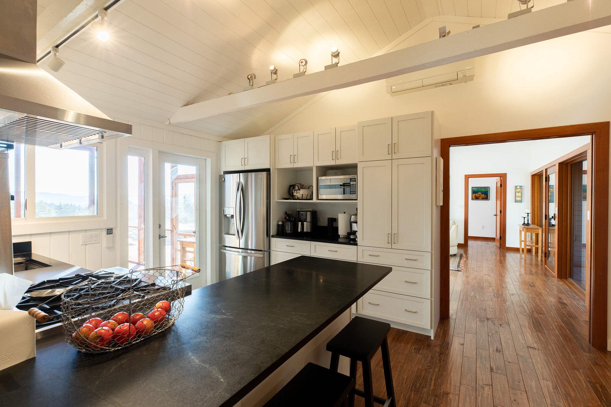 A modern kitchen with sleek white cabinetry, dark countertops, a stainless steel refrigerator and microwave, and a central island with bar stools, offering access to an outdoor deck.