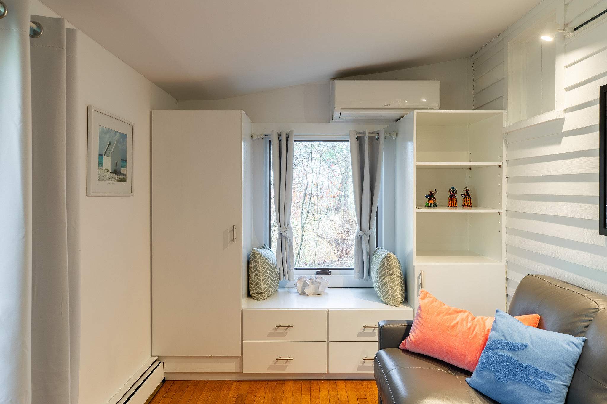 A cozy sitting area with a built-in window seat flanked by white storage cabinets, a comfortable grey armchair with colorful pillows, and a large window overlooking trees.