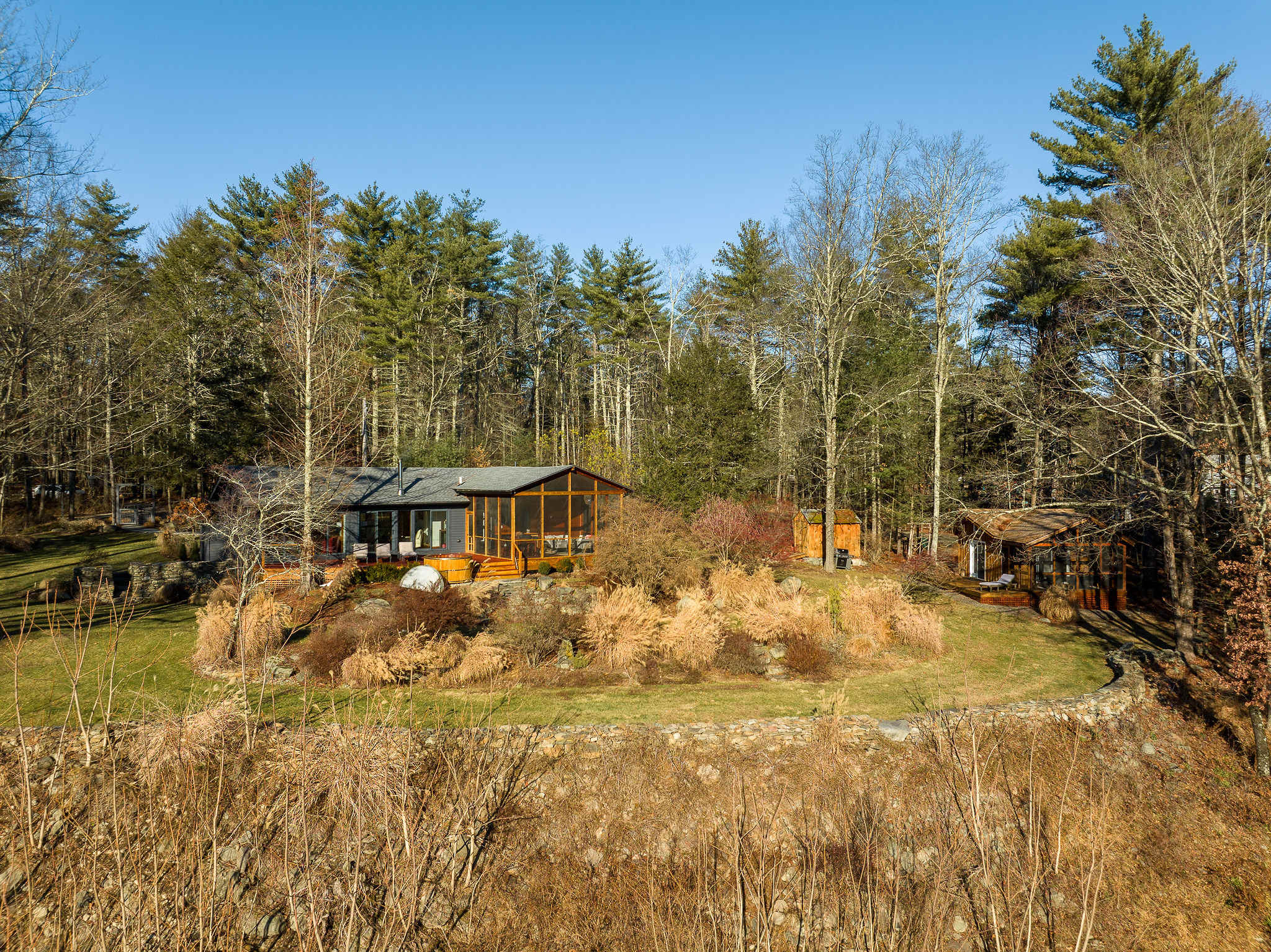 An expansive aerial view showcases a modern house with a large wooden deck, hot tub, and screened porch, nestled amidst a vast wooded landscape with distant mountains. The property features a well-maintained lawn and natural rock walls.