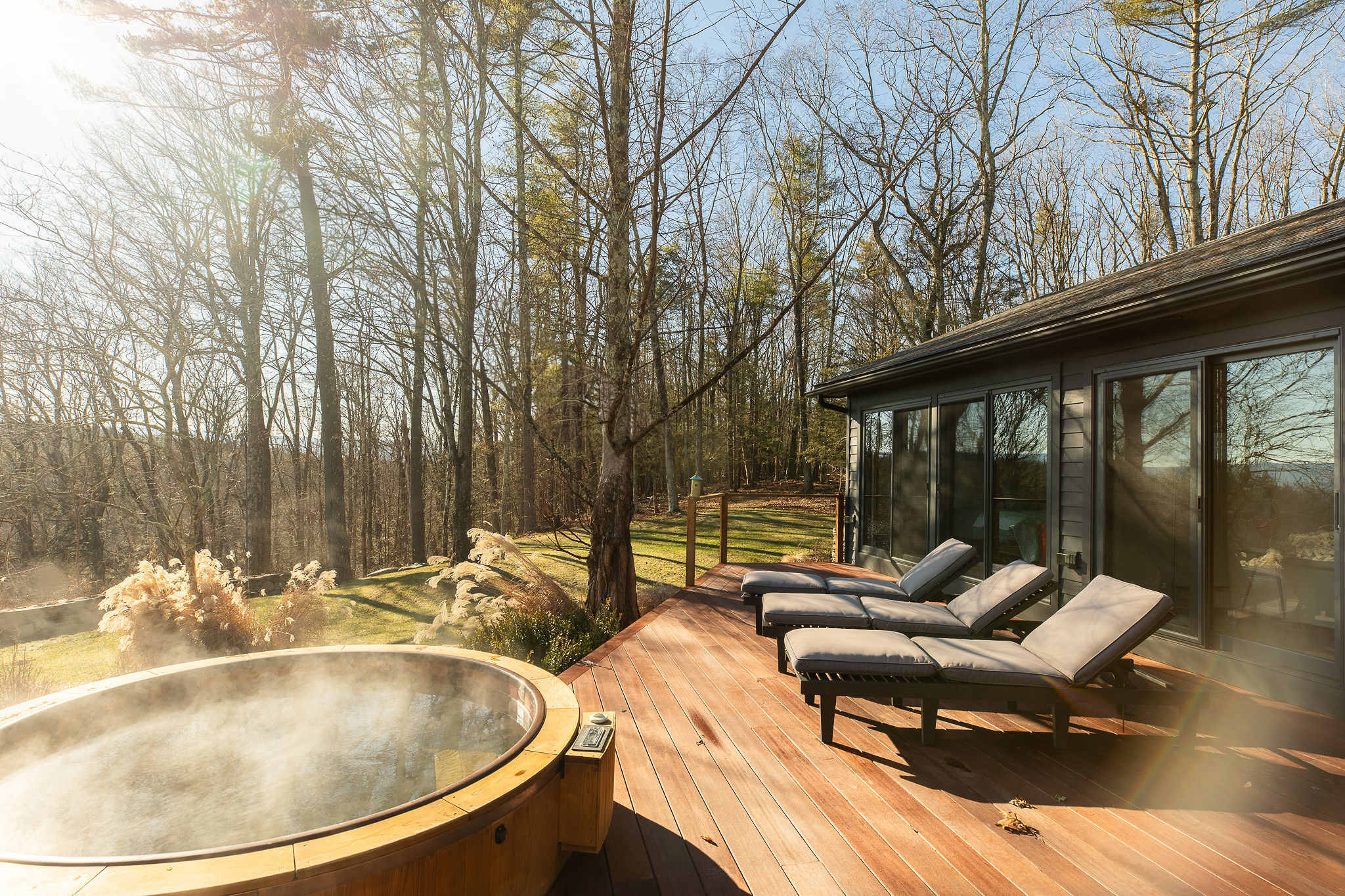 A spacious wooden deck with a steaming round hot tub and three grey lounge chairs extends from a modern house with large glass sliding doors. The property is surrounded by tall, bare trees under a bright, sunny sky, offering a serene natural setting.