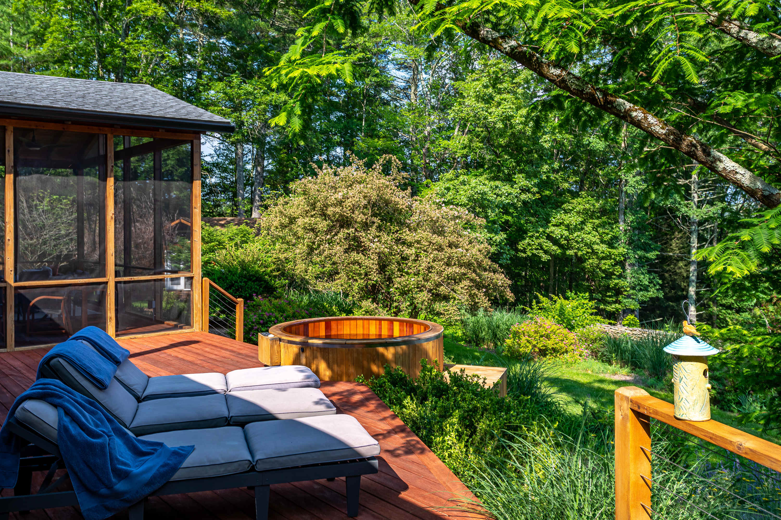 A wide wooden deck features two grey lounge chairs with blue blankets, overlooking a round wooden hot tub and a lush green backyard with dense trees. A screened porch is visible to the left, offering additional outdoor living space.