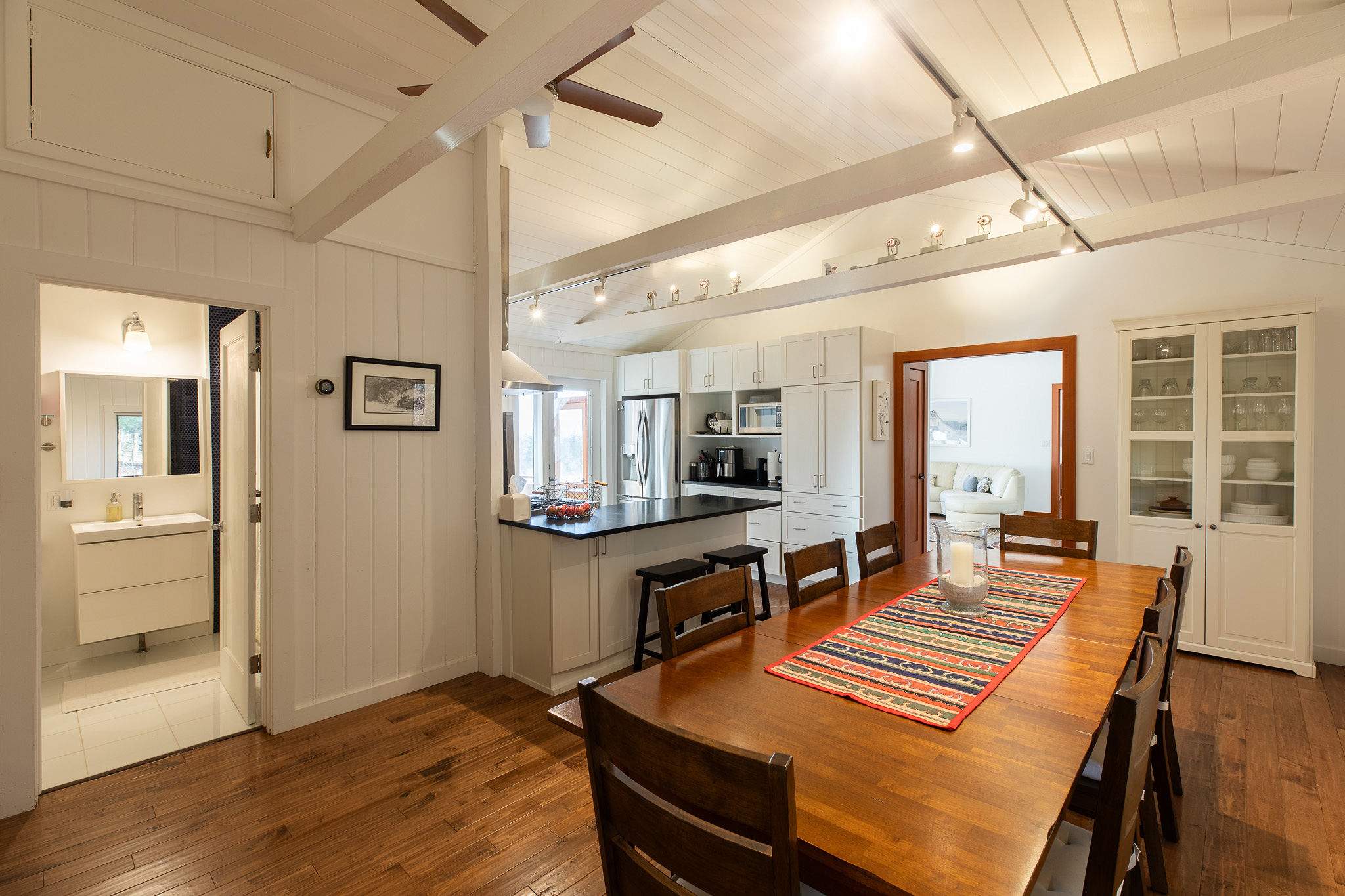 An open-concept dining area with a long wooden table and eight chairs, featuring exposed white ceiling beams, track lighting, and hardwood floors, adjacent to a kitchen and a doorway to another room.