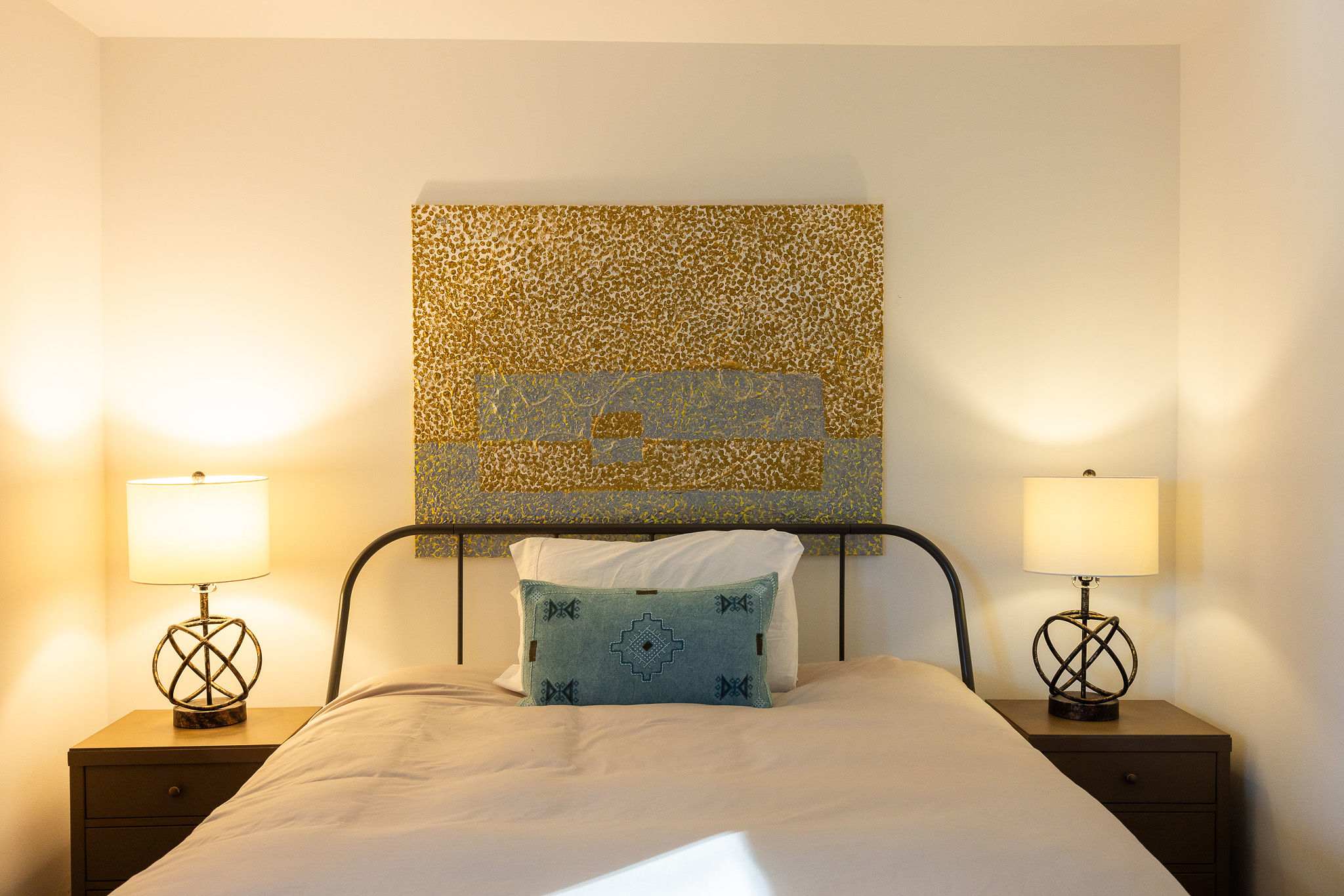 A close-up view of a queen-sized bed with a black metal frame, white bedding, and a blue patterned accent pillow, flanked by two dark wood bedside tables with lamps and a large abstract artwork above.