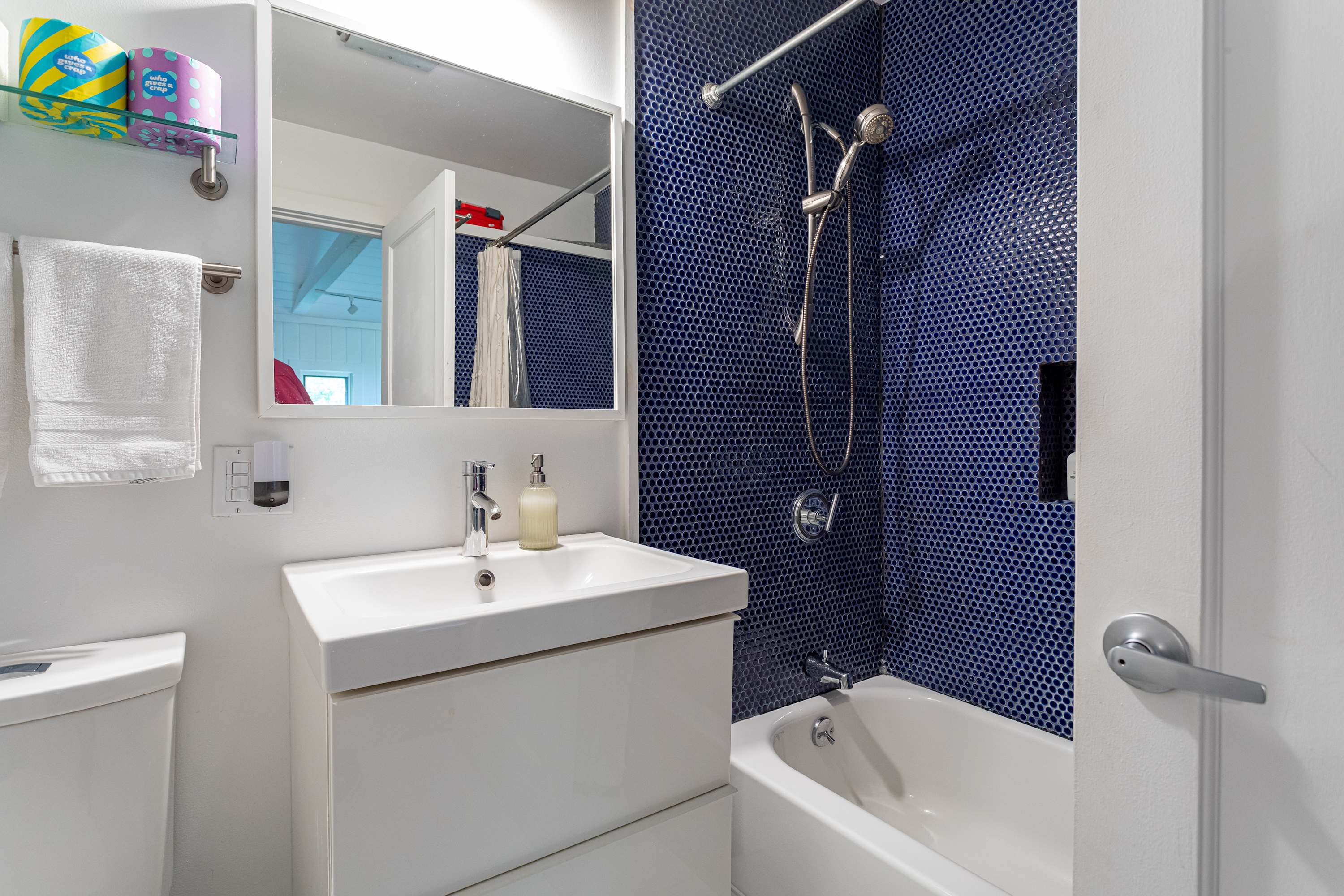 A compact bathroom featuring a white floating vanity with a sink, a toilet, and a bathtub with a shower enclosed by vibrant blue penny tile walls.
