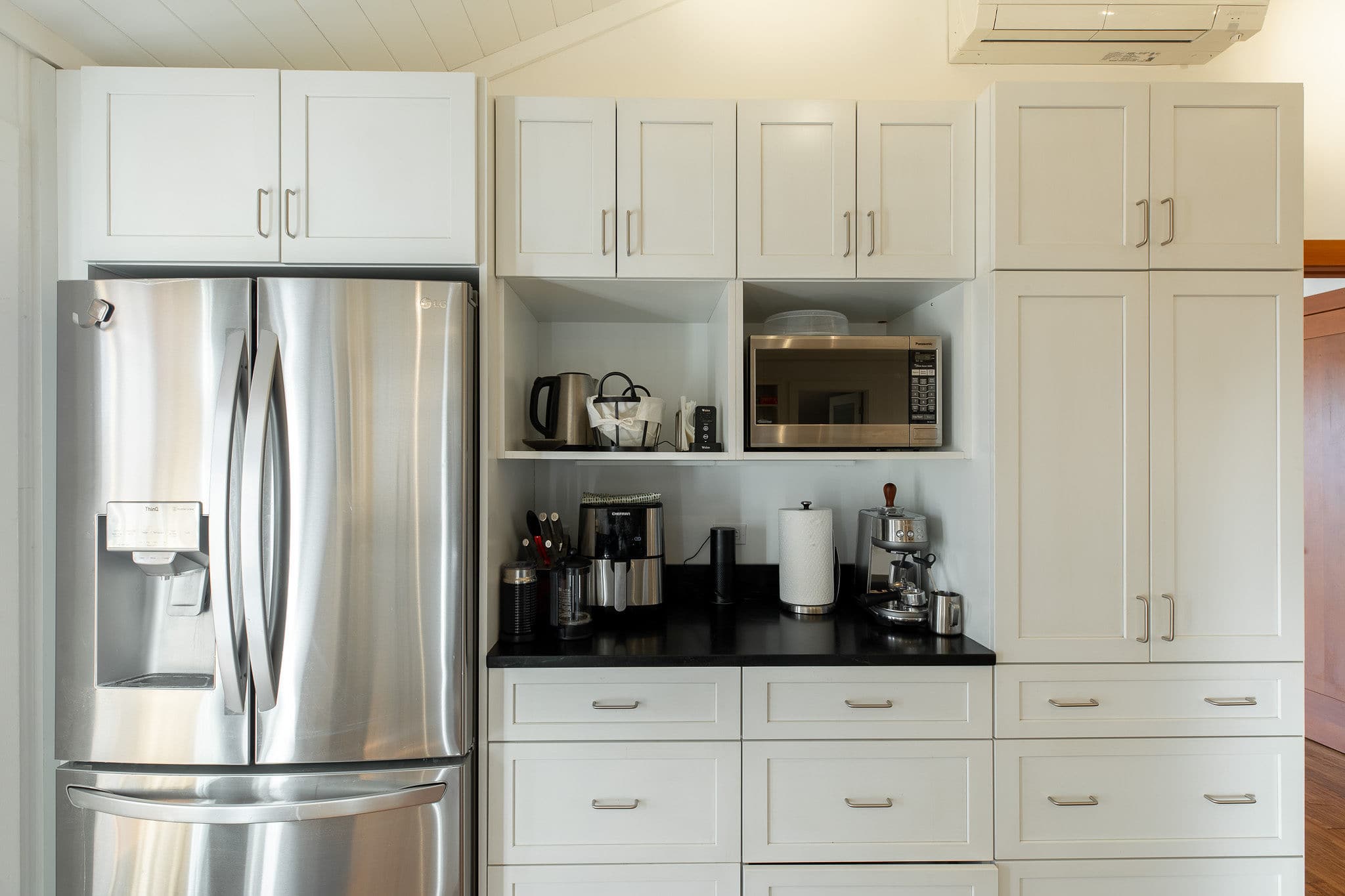 A bright, modern kitchen featuring white shaker-style cabinets, a large stainless steel French door refrigerator, and black countertops with various coffee-making appliances and open shelving.