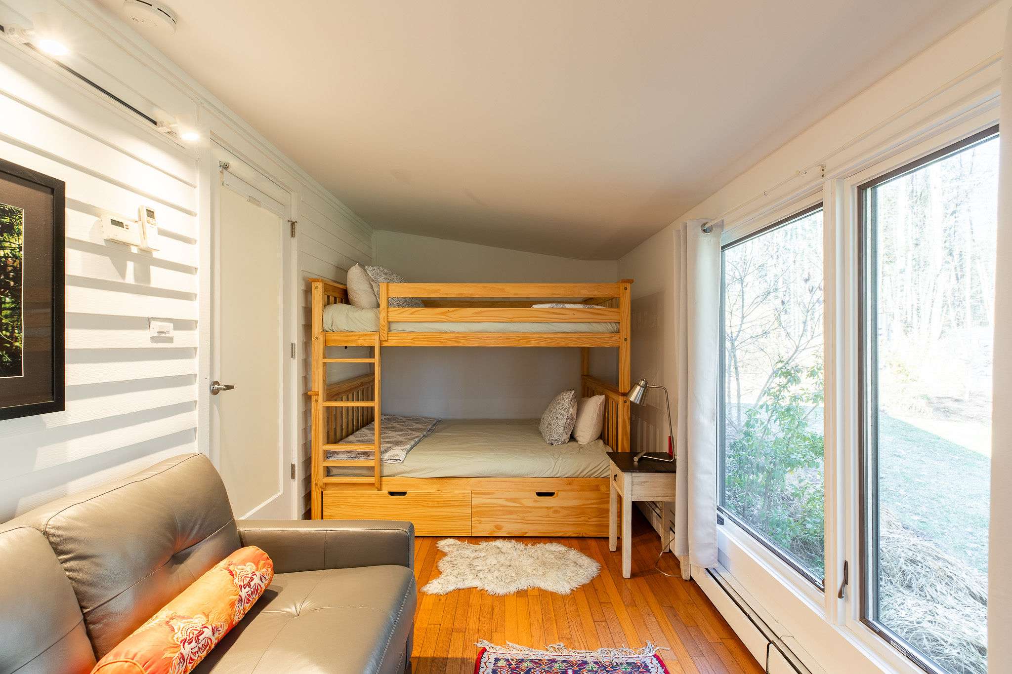 A bright bedroom featuring a light wood bunk bed with a trundle drawer, a small grey sofa, and large windows providing ample natural light and a view of the outdoors.