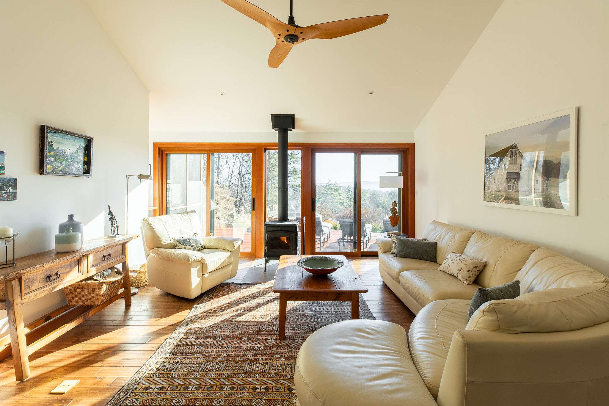 Bright living room featuring a light leather sectional sofa, a wooden coffee table, and a wood-burning stove, with a wooden console table on the left and large glass doors offering outdoor views.