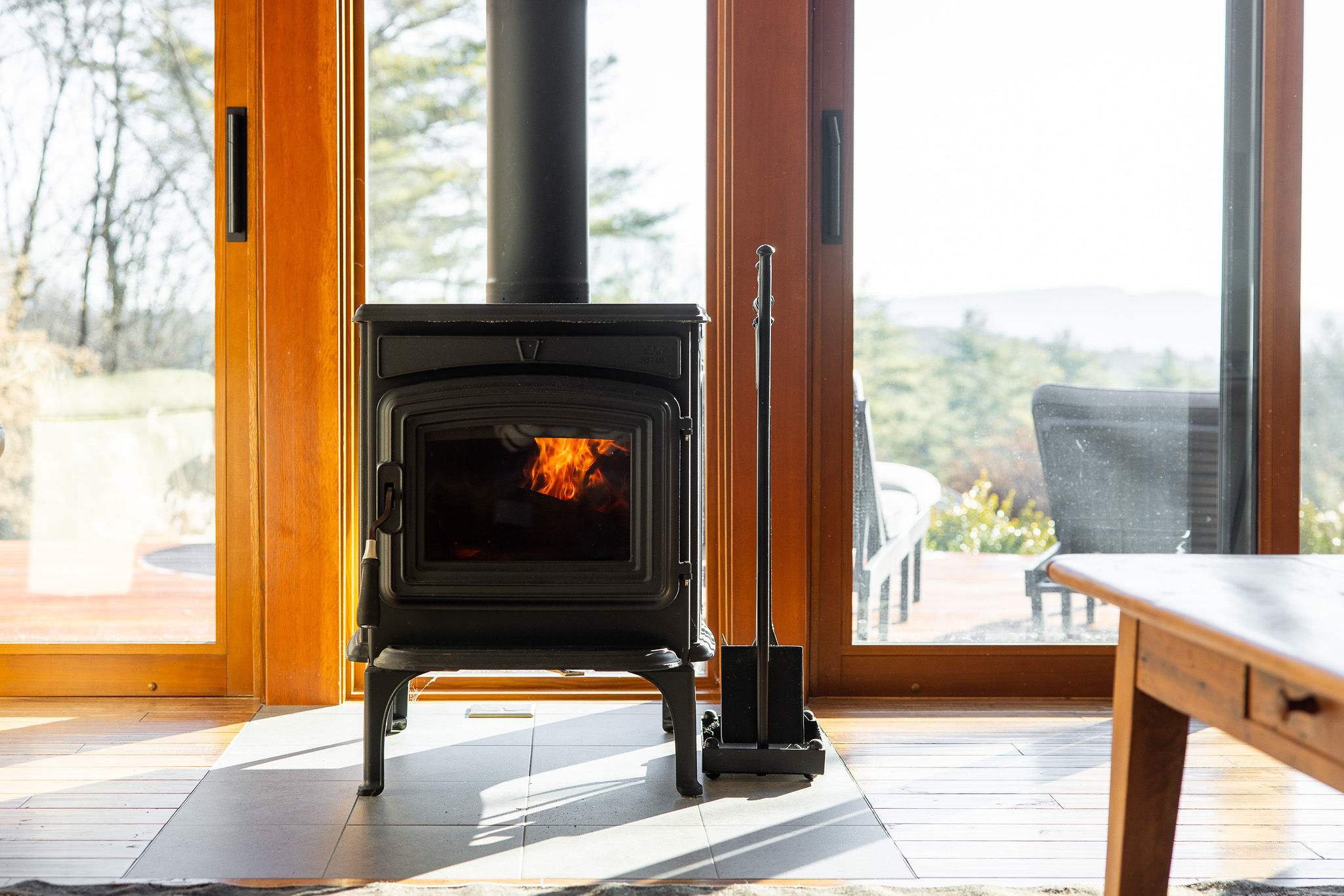 Close-up of a black cast-iron wood-burning stove with visible flames, set on a tiled hearth between large glass sliding doors that offer a glimpse of the outdoor deck.