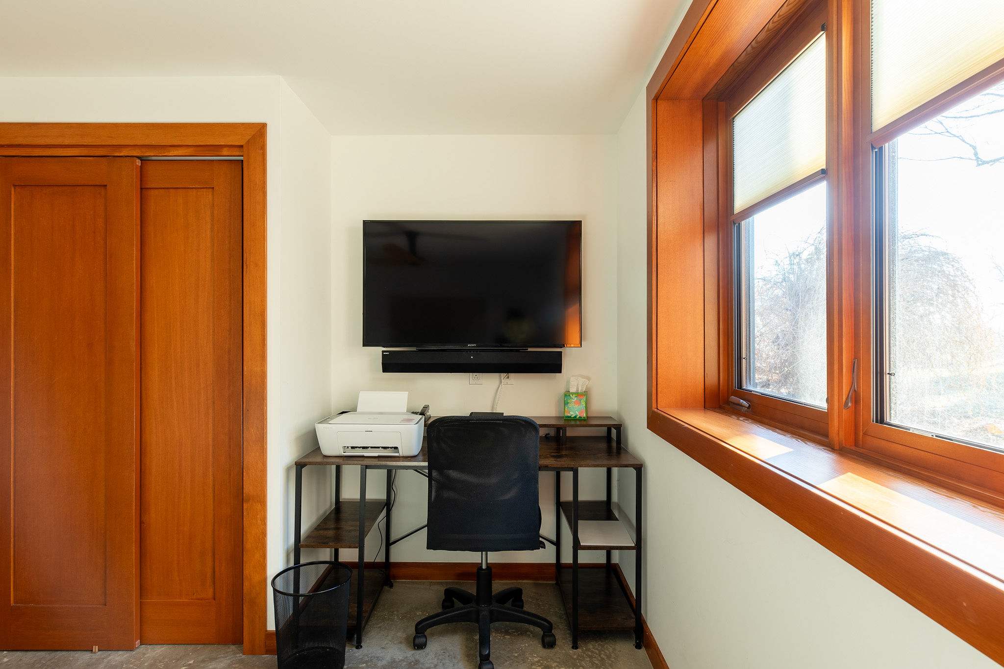 A dedicated workspace featuring a dark metal and wood desk with a printer and an office chair, positioned beneath a wall-mounted TV and next to a large window with warm wooden trim.