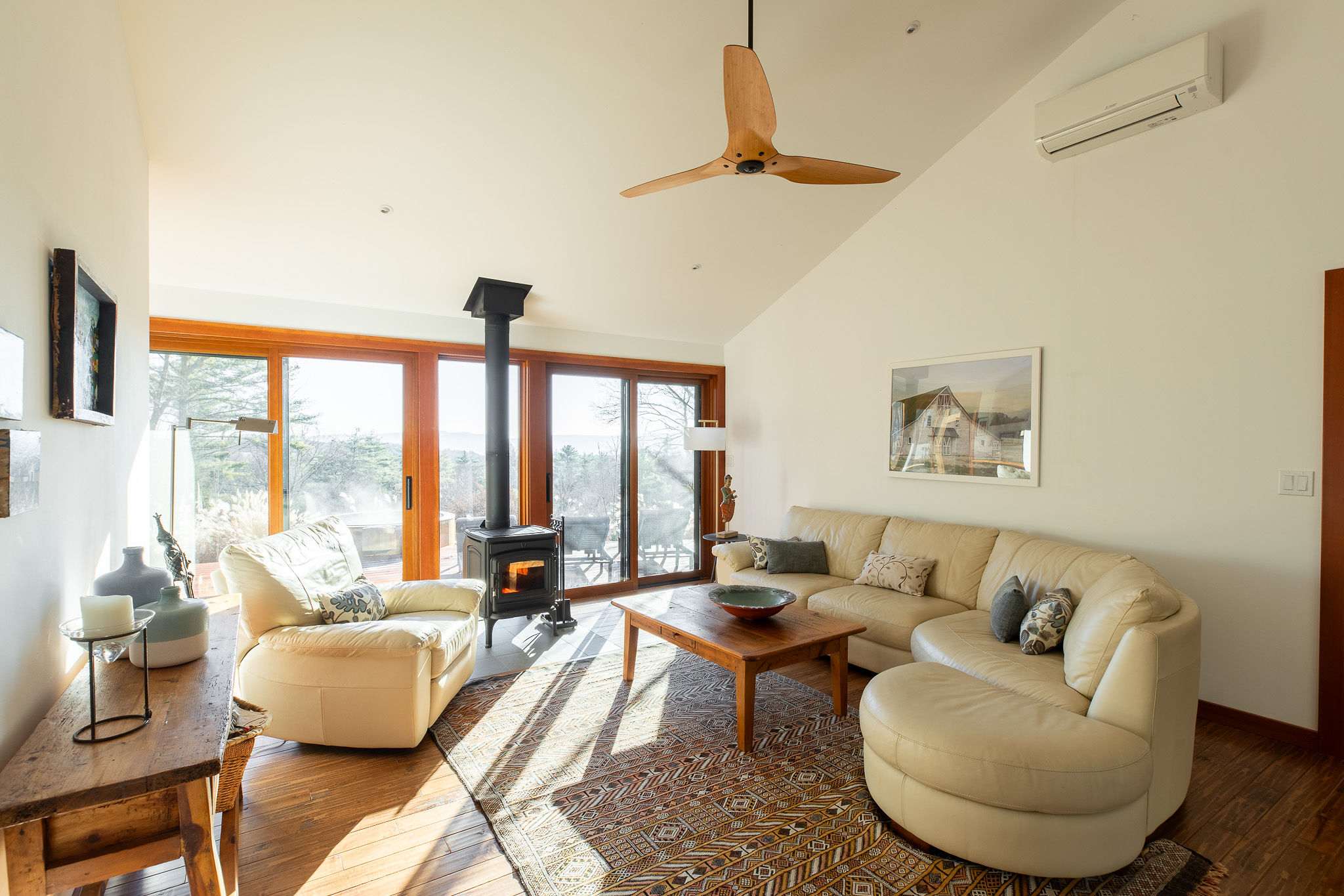 Sunlit living room with vaulted white ceilings, a large light leather sectional sofa, a wooden coffee table, and a wood-burning stove, with expansive views through glass doors.