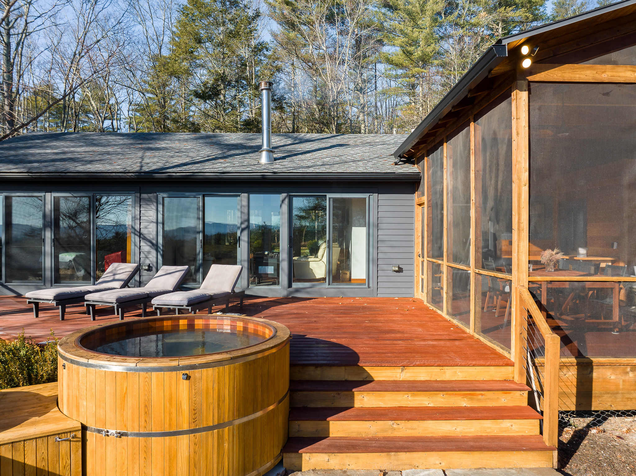 The exterior of a modern house features a large wooden deck with a round hot tub and lounge chairs, adjacent to a screened-in porch. The house has dark siding and large windows, set against a backdrop of tall trees and a clear blue sky.