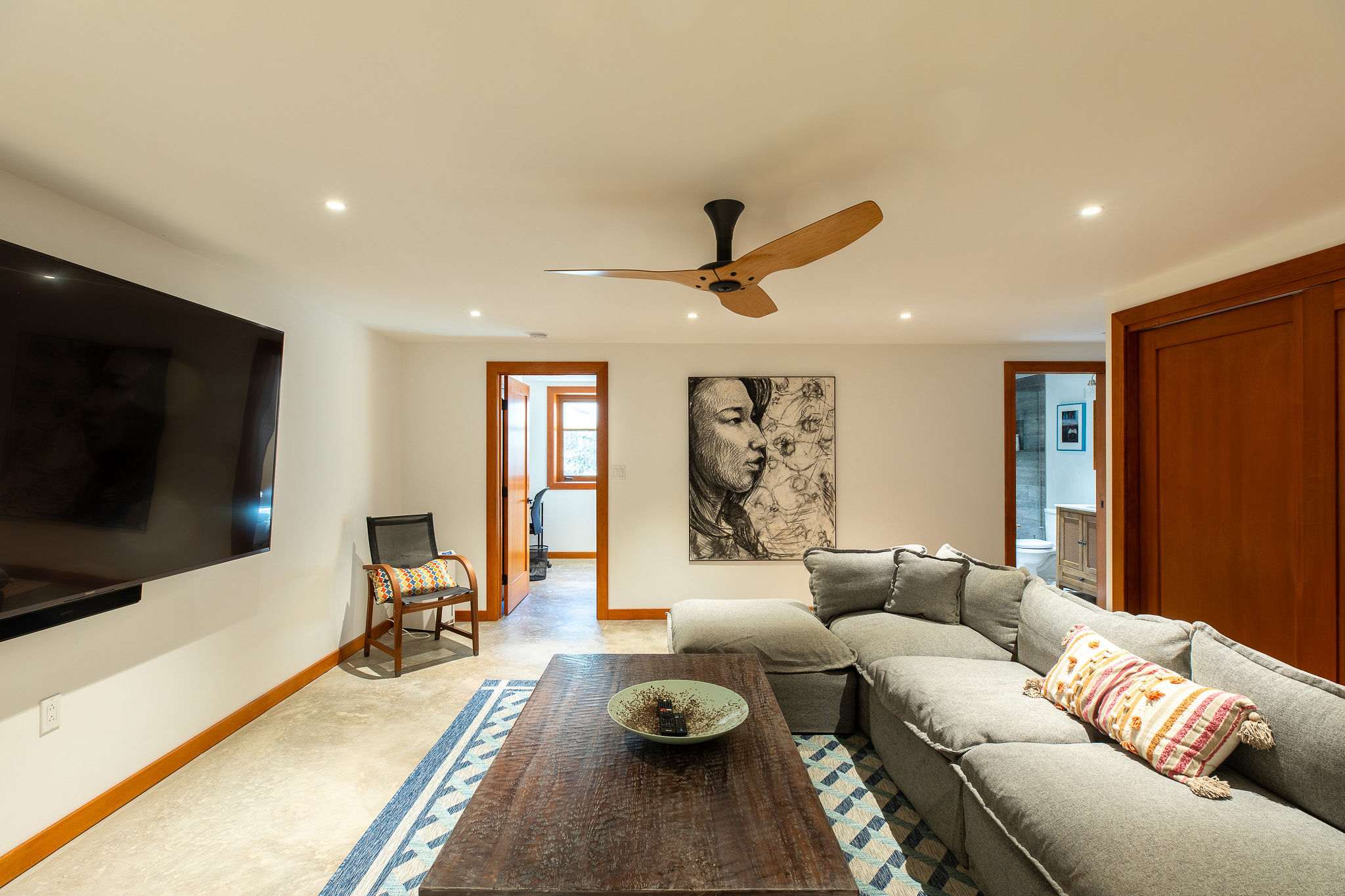 A spacious main living room with a large grey sectional sofa, a rustic wooden coffee table, a wall-mounted television, and a modern wooden ceiling fan, with doorways leading to other areas.
