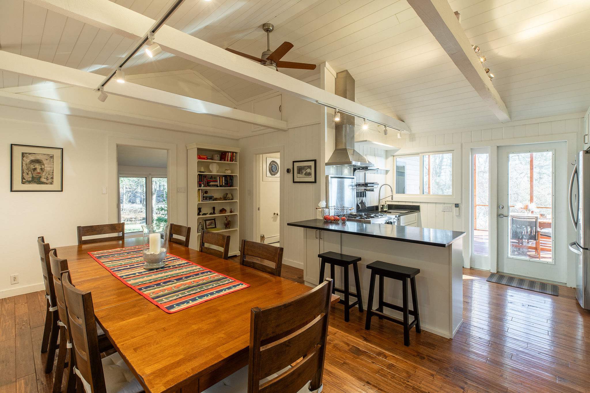 An open-concept dining and kitchen area featuring a long wooden dining table, a central kitchen island with bar stools, and stainless steel appliances, all under a vaulted white ceiling with exposed beams.