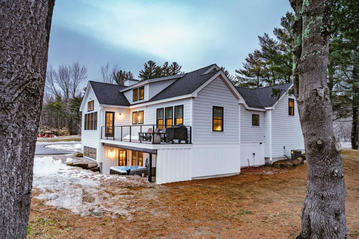 The back of a modern white farmhouse with a black roof and large windows, featuring a deck with a black railing and a hot tub on the lower level. Patches of snow and bare ground are visible, with tall trees in the background under a cloudy sky.