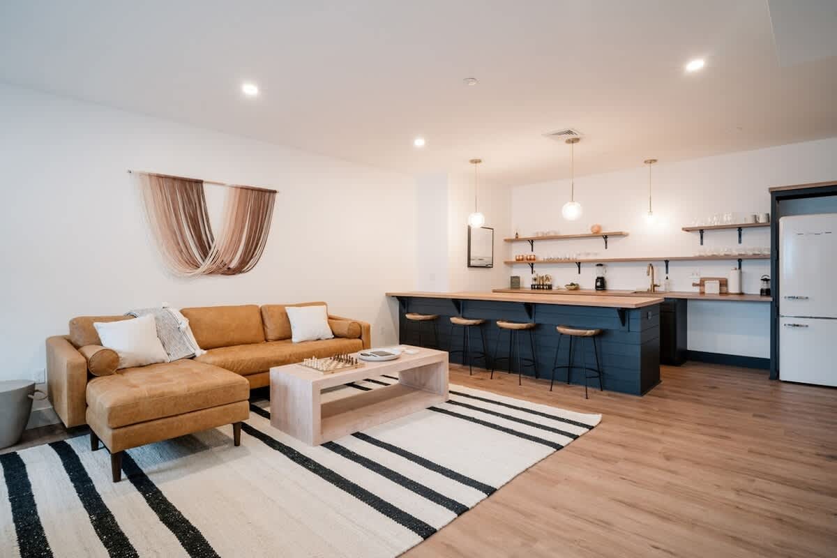 Bright basement living area featuring a comfortable brown sectional sofa, a light wood coffee table, a striped rug, and a dark blue bar with wooden countertops and bar stools, creating a relaxed entertainment space.