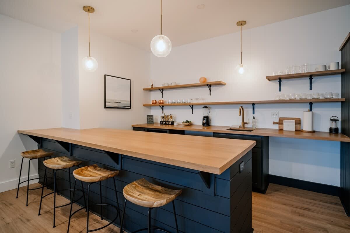 A stylish wet bar features a long wooden countertop, dark blue shiplap-style base cabinets, and open wooden shelving displaying glassware. Three clear globe pendant lights illuminate the space, complemented by modern bar stools.