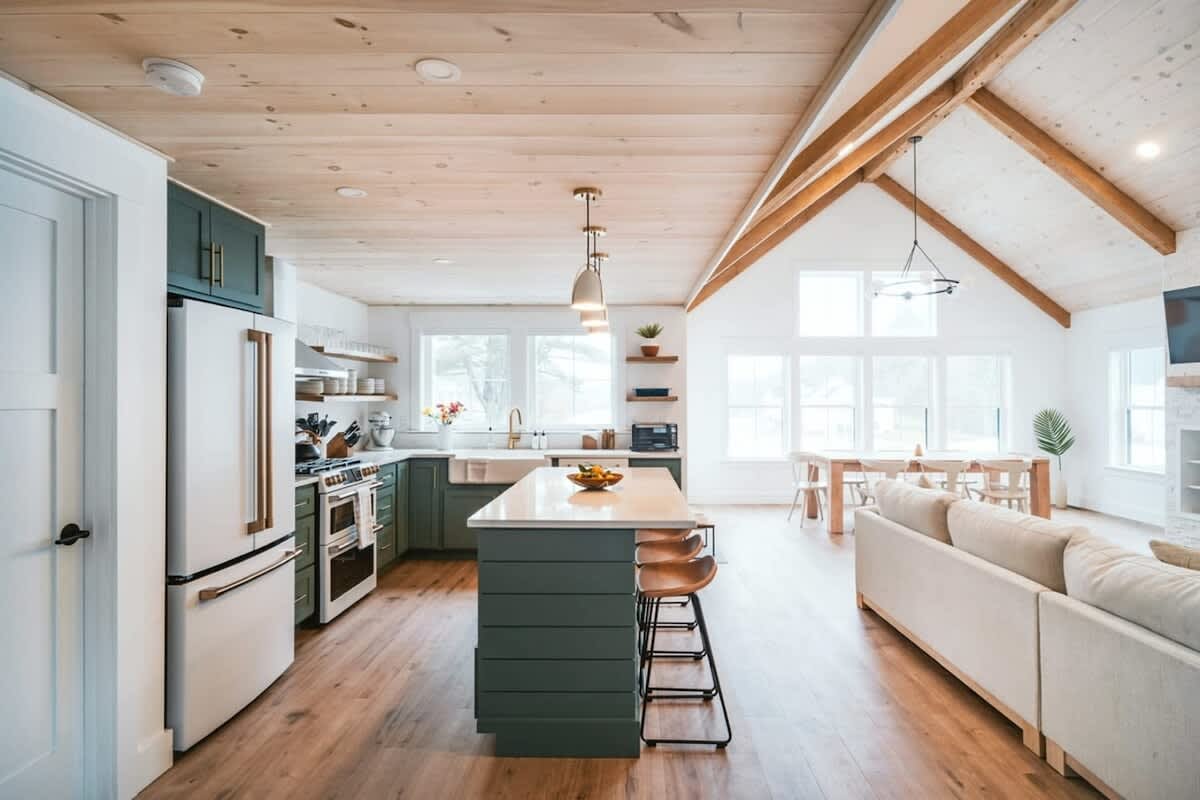 Expansive open-concept kitchen, dining, and living area with vaulted wooden ceilings and exposed beams. The kitchen features a large island with bar seating, dark green cabinets, and modern appliances, flowing into the dining and living spaces.