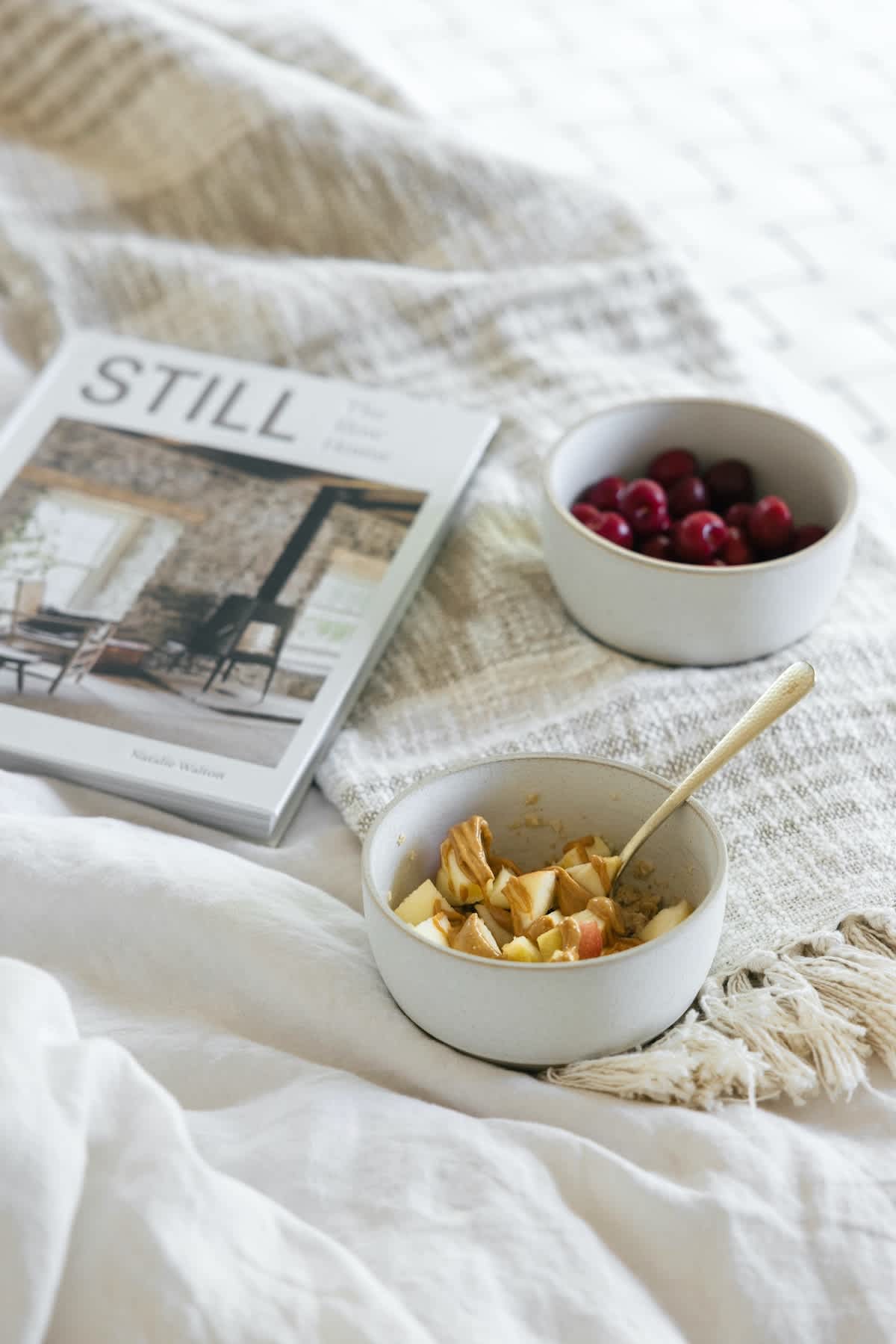 Cozy breakfast scene with a bowl of oatmeal with apples and peanut butter, a bowl of cherries, and a book on a white bed with a textured throw blanket.