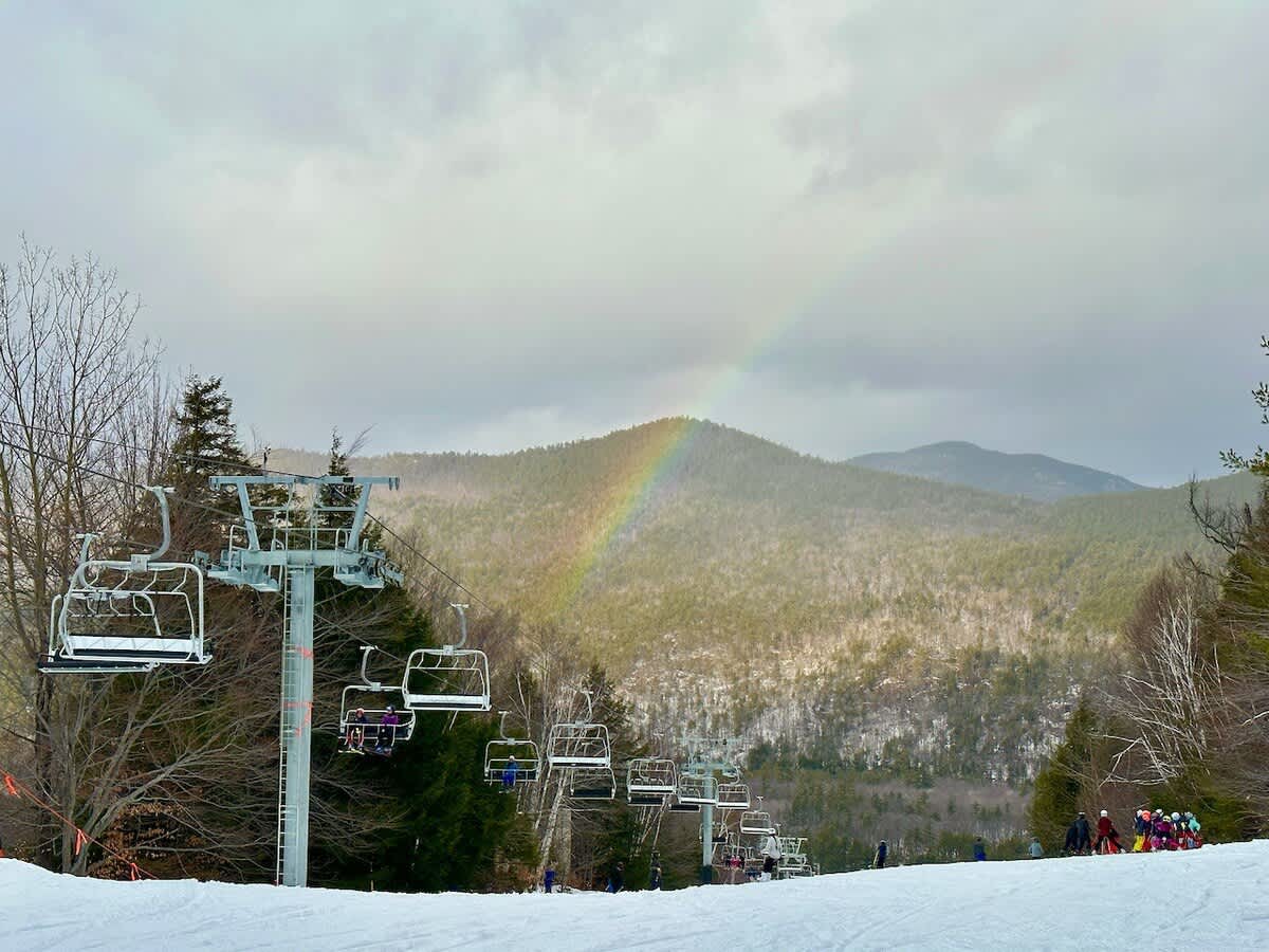 A vibrant ski slope scene with multiple chairlifts ascending a snowy mountain, a colorful rainbow arching over a tree-covered peak, and skiers gathered at the base under a cloudy sky.