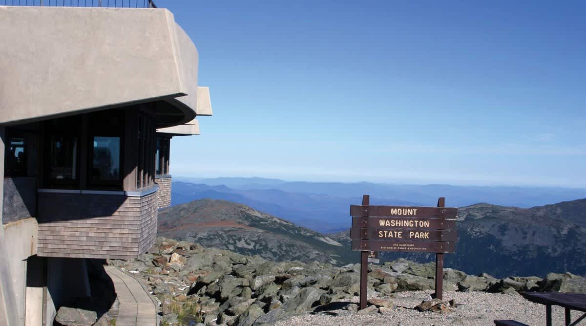 A rugged mountain landscape featuring a modern stone and wood building on a rocky summit, with a 'Mount Washington State Park' sign and vast mountain ranges stretching into the hazy distance under a clear blue sky.