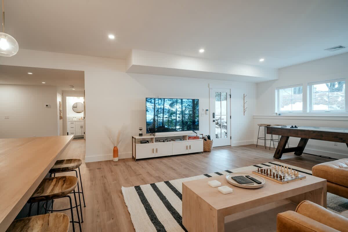 Expansive basement game room and living area with a large TV entertainment center, a pool table, and a bar area, all set on light wood flooring with ample natural light from multiple windows.