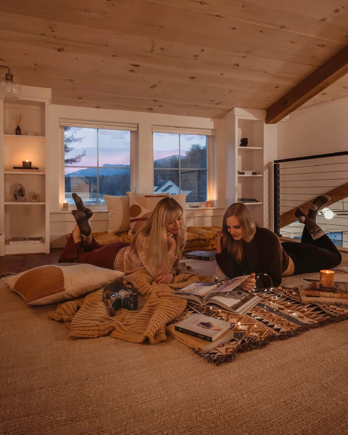 Cozy loft space with a sloped wooden ceiling, featuring two women relaxing on the floor with books and blankets, illuminated by string lights. Large windows offer a view of the outdoors at dusk.
