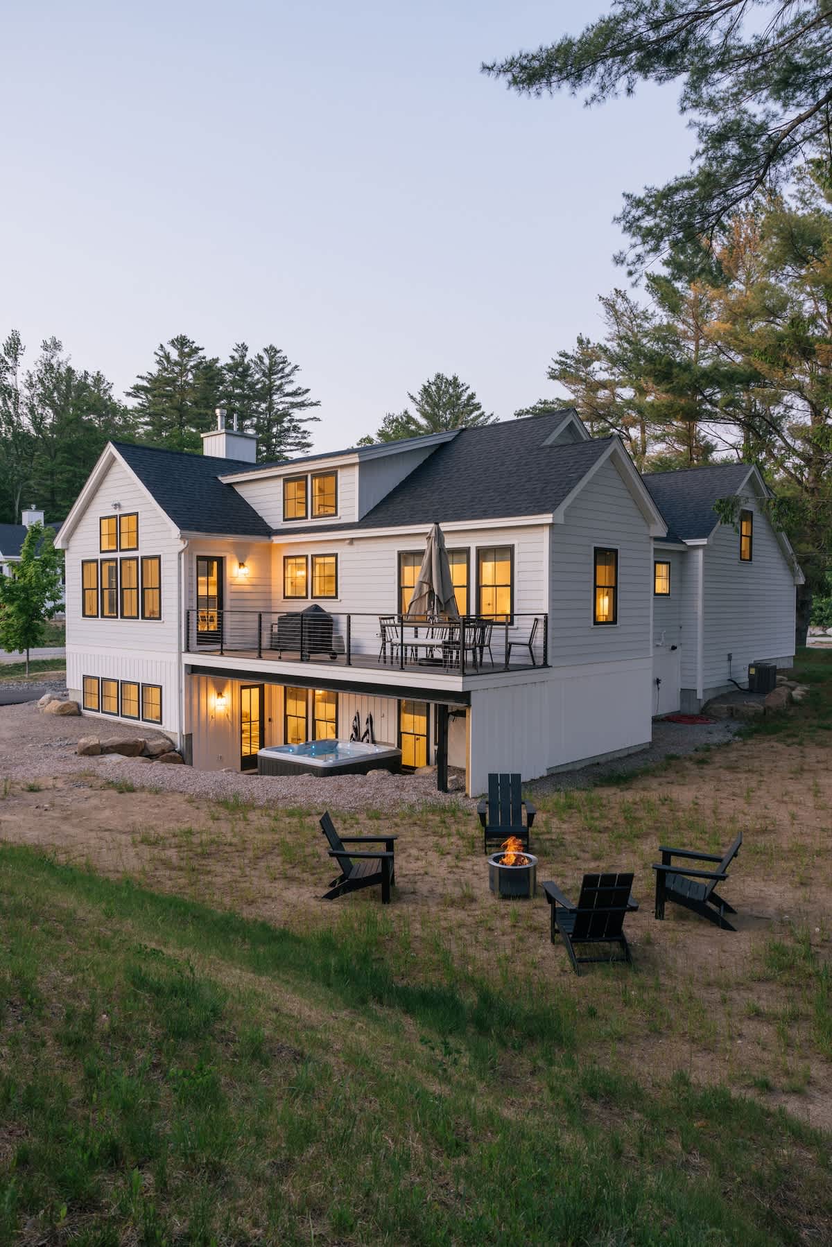 Modern farmhouse exterior with white siding and black-framed windows, featuring a spacious back deck with outdoor seating and a hot tub on the lower level, surrounded by a grassy yard and trees.