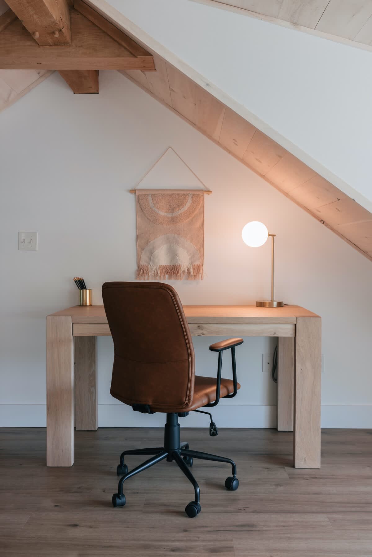 Dedicated workspace in a loft with a sloped wooden ceiling, featuring a light wood desk, a brown leather office chair, and a modern globe lamp. A woven wall hanging adds a touch of decor.
