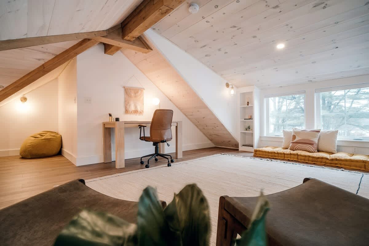 Bright loft area with a sloped wooden ceiling and exposed beams, featuring a light wood desk with a brown leather office chair and a decorative wall hanging. A yellow bean bag chair is in the corner.