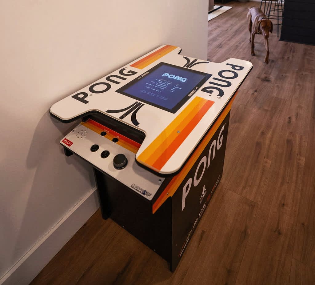 A vintage-style Pong arcade game machine with a classic Atari design stands on hardwood floors. The screen displays the game, and control knobs are visible, offering a nostalgic gaming experience.