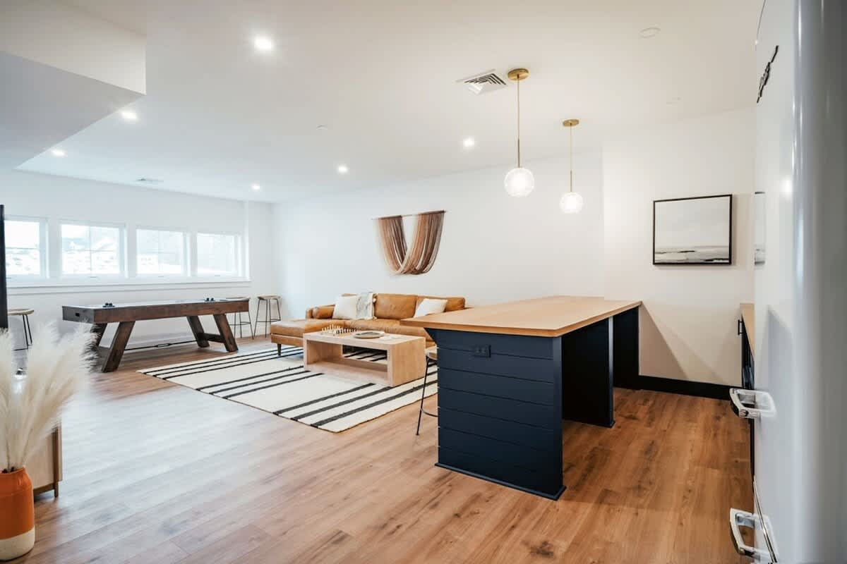 Wide view of a versatile basement living and game room, showcasing a brown sectional sofa, a pool table, a dark blue bar with seating, and a striped area rug, designed for entertainment and relaxation.