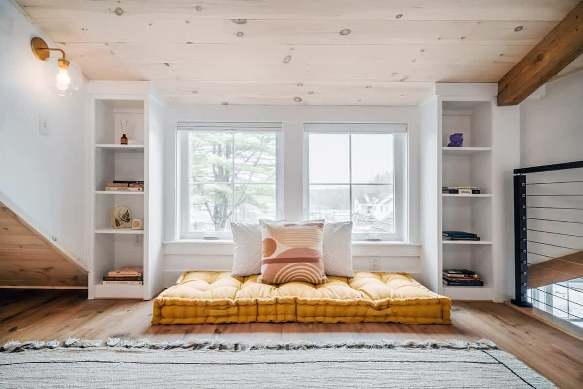 Bright loft space with a sloped wooden ceiling and built-in shelves flanking large windows, featuring a long yellow floor cushion with decorative pillows. The room has light wood flooring and a textured rug.