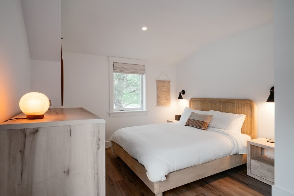 Bright bedroom with a light wood bed frame and rattan headboard, white bedding, a light-colored dresser with a spherical lamp, and a window with a Roman shade, all set on light wood flooring.