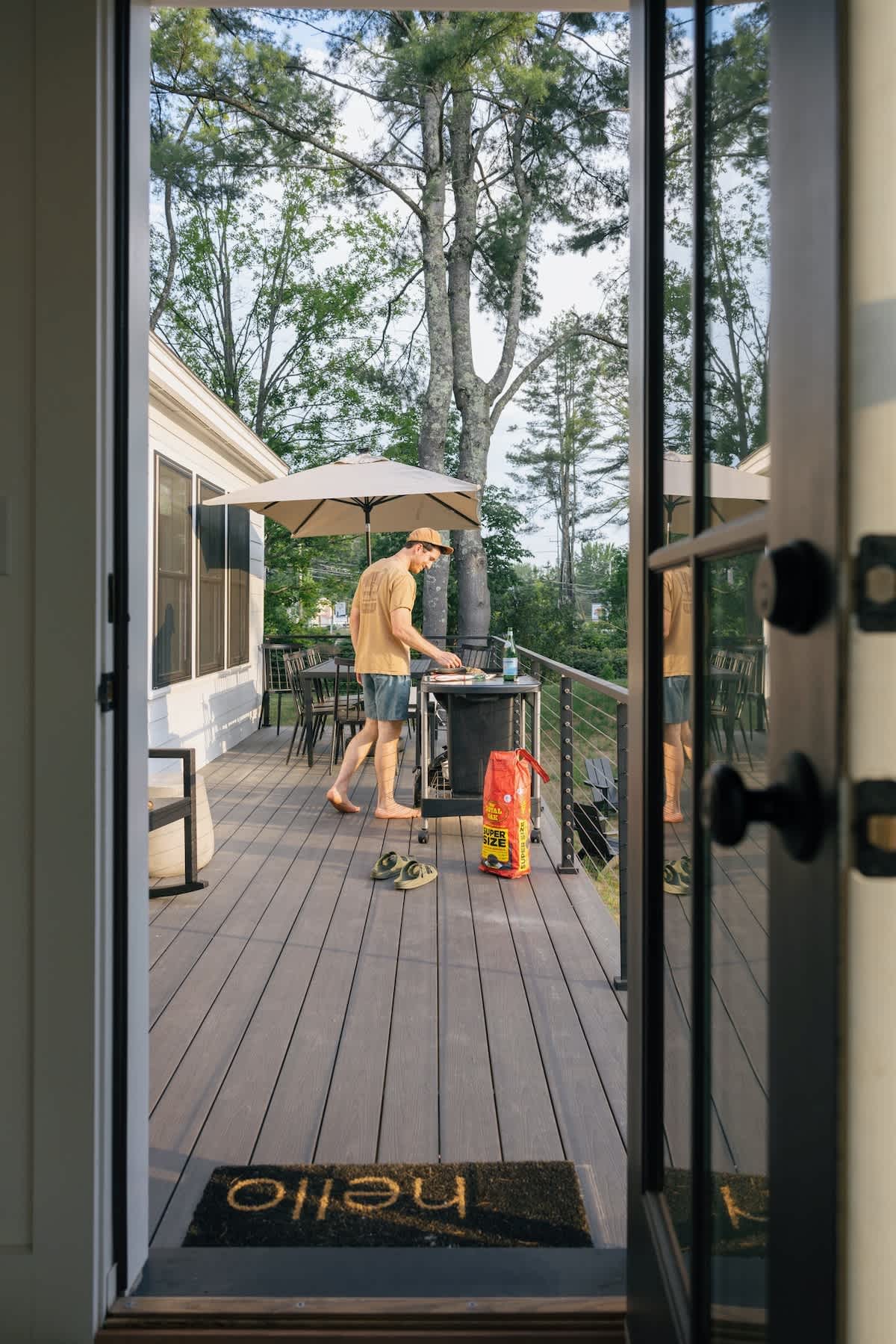 A man in a t-shirt and shorts is grilling on a dark gray deck, viewed from an open doorway with a 'hello' mat. Tall trees are visible in the background, and a beige umbrella shades the outdoor dining area.