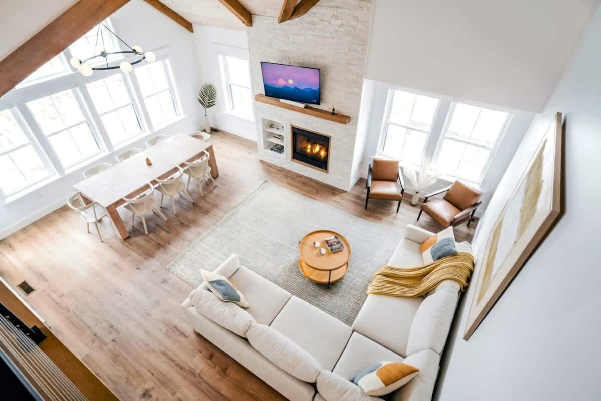 Spacious open-concept living and dining area with vaulted wooden ceilings and exposed beams. A large sectional sofa faces a white brick fireplace with a mounted TV, adjacent to a long dining table with modern chairs.