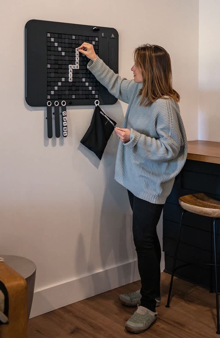 A woman stands next to a wall-mounted, oversized Scrabble-style game board, placing a letter tile. The black board features white letter tiles and small bags for game pieces, adding a unique entertainment option.